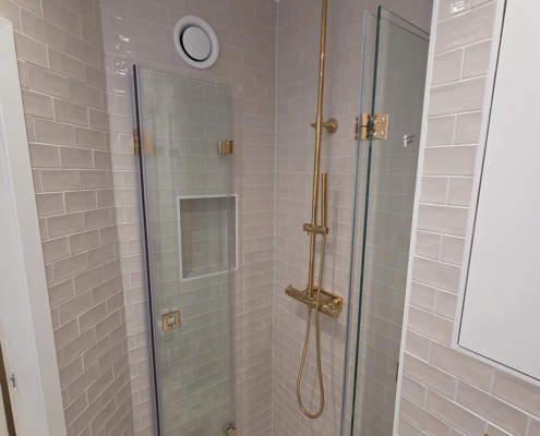 Hörndusch med guldarmatur och regnduschhuvud, glasdörrar och infälld nisch i beiga kakelvägg – mosaik på golvet med guldgaller.
