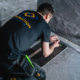 En hantverkare från Grand Construction Stockholm AB knäböjer och installerar ett golvbrunnsram i ett badrum med mörkgrå marmorklinker under en badrumsrenovering i Stockholm.