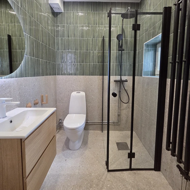 Badrumsrenovering i Stockholm med grönt vertikalt kakel, grå natursten, svarta armaturer och glasdusch – utfört av Grand Construction